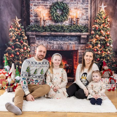 Christmas Photography A family of four sits on a rug in front of a festive Christmas backdrop, decorated trees, and gifts.